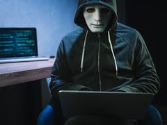 一个戴着面具和兜帽的人在昏暗的房间里使用笔记本电脑。