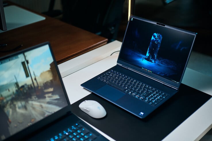 一台深蓝色中端游戏笔记本电脑（Alienware Aurora 16X）斜靠在桌垫上，桌垫上显示着一幅发着蓝光的科幻壁纸；电脑旁边放着一只白色无线鼠标，前景中还有另一台模糊的笔记本电脑。