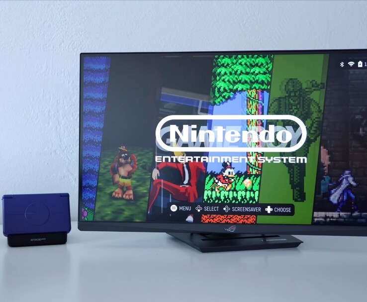 紫色 Retroid Pocket Flip 2 放在显示器旁边的底座上，电视屏幕上显示复古 NES 模拟器界面。(图片来源：YouTube 上的 Retro Game Corps）