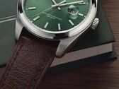 Timex Legacy Day and Date 腕表特写，展示其绿色太阳纹表盘、银色表圈和拉丝表耳、带凹槽的表冠以及纹理棕色皮革表带，背景为深绿色盒子和木质表面。