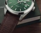 Timex Legacy Day and Date 腕表特写,展示其绿色太阳纹表盘、银色表圈和拉丝表耳、带凹槽的表冠以及纹理棕色皮革表带,背景为深绿色盒子和木质表面。