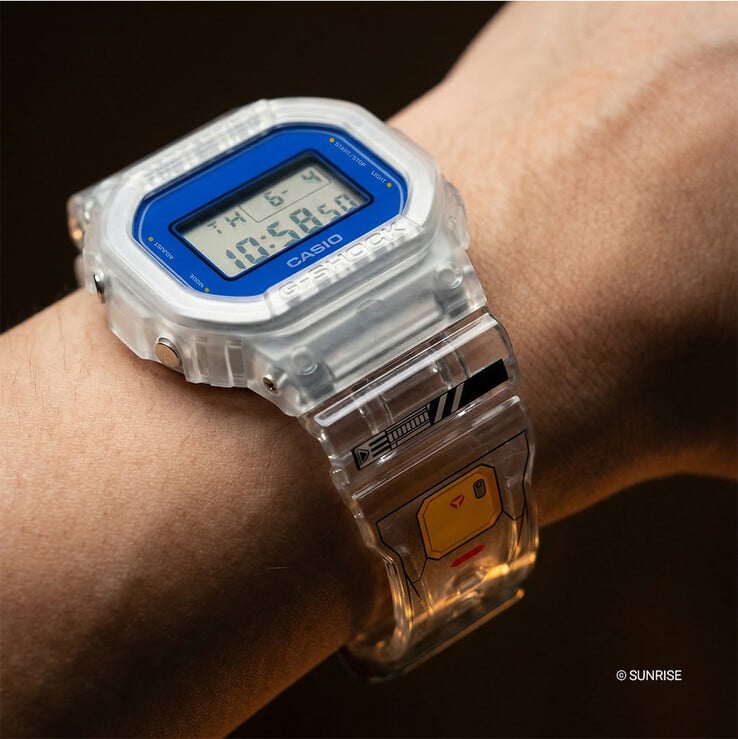 佩戴在手腕上的卡西欧 G-Shock x 机动战士高达 DW-5600GM26-7DR，采用透明树脂表壳和表带，蓝色表圈环绕，表带上印有以高达为灵感的图案。