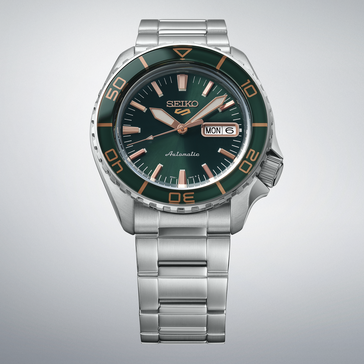 Seiko 5 Sports SKX HDB005 配有深绿色表盘、同色绿色表圈、玫瑰金色指针和时标以及不锈钢表链,展示于浅灰色背景前。