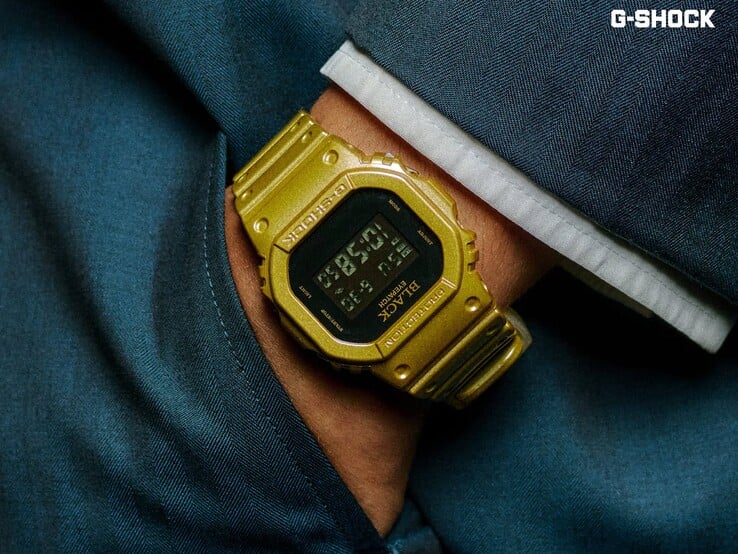 一张特写照片,展示一款金色卡西欧G-Shock DW-5600BEP-9手表戴在手腕上,背景为深蓝色布料。该手表配有黑色数字显示屏、金色表壳和表带,表圈和表盘上印有BlackEyePatch品牌标识。