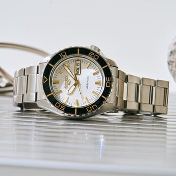 Seiko 5 Sports SKX HDB004 腕表,配备银白色表盘、带金色刻度的黑色旋转表圈、金色指针和时标,以及不锈钢表链,以倾斜角度拍摄。