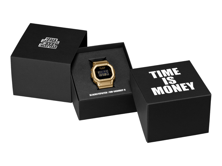 一款金色卡西欧G-Shock DW-5600BEP-9手表,配有黑色数字显示屏,置于两个黑色展示盒之间。表盘上印有BlackEyePatch品牌标识,而前景中的盒子上则印着“TIME IS MONEY”字样。