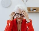 一位白发、戴着眼镜的年长女性,坐在一间光线明亮的客厅里,一边打电话,一边用手捂着太阳穴。