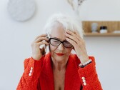 一位白发、戴着眼镜的年长女性，坐在一间光线明亮的客厅里，一边打电话，一边用手捂着太阳穴。