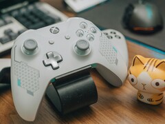 白色的 Xbox 控制器放在木质书桌上，旁边摆着一只小猫公仔（图片来源：FOX ^.ᆽ.^= ∫ via Pexels）