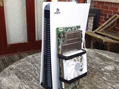 一台 PlayStation 5 游戏机放置在一块定制的 AMD BC250 电路板旁边，该电路板安装有散热片和冷却风扇。(图片来源：Budget-Builds Official via YouTube）